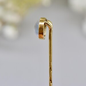 May include: A gold-colored stick pin with a round, light-colored stone at the top. The pin has a simple, elegant design, with a slightly curved top and a textured shaft. The stone appears to be a moonstone or similar iridescent gem.
