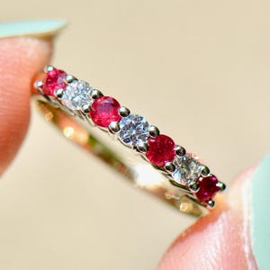 May include: A gold ring with a row of alternating red and clear gemstones. The gemstones are round and set close together, creating a continuous line of color. The ring is held between two fingers.