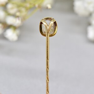 May include: A close-up of a gold-colored stick pin. The pin features a square-shaped, light-colored stone set in a gold bezel. The pin's stem is long and has a twisted design.