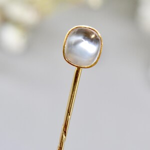 May include: A gold-colored hairpin with a square, clear, iridescent stone set in a gold bezel. The pin has a twisted design. The background is blurred, with soft white and gray tones.