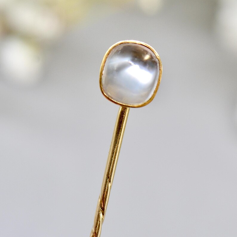 May include: A gold-colored hairpin with a square, clear, iridescent stone set in a gold bezel. The pin has a twisted design. The background is blurred, with soft white and gray tones.