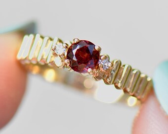 May include: A gold ring with a red gemstone and two smaller white gemstones. The ring has a textured band.