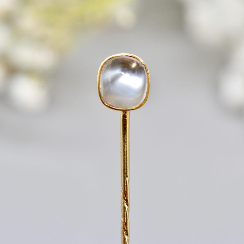 May include: A close-up of a gold-colored hat pin with a square, iridescent, off-white stone set in a gold bezel. The pin's stem is twisted, and the background is blurred.