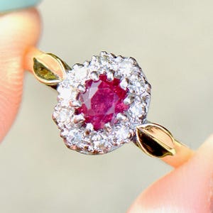 May include: A gold ring featuring a central, round, deep red ruby surrounded by a halo of small, clear diamonds. The ring's band has a delicate design with leaf-shaped accents on either side of the gemstone setting.