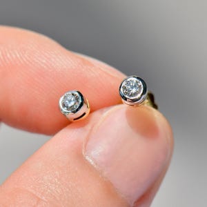 Puede incluir: Un par de pendientes de botón con piedras redondas, transparentes y facetadas, engastadas en un bisel de tono plateado pulido. Cada piedra está asegurada dentro de un marco circular de metal, mostrando un corte brillante y una superficie reflectante.