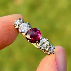 May include: A gold ring with a central, round, deep red ruby, and four smaller, round, clear diamonds. The ring is held up against a blurred green background, highlighting the gemstones' sparkle.