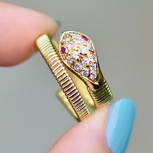 以下が含まれることがあります： ルビーとダイヤモンドの頭を持つ金の蛇の指輪。指輪は青いマニキュアを塗った指にはめられています。