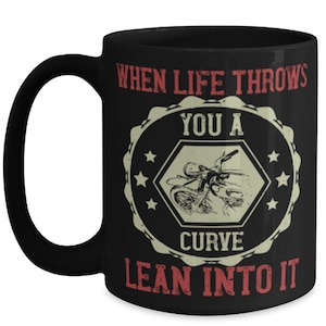 May include: Black ceramic coffee mug with a white and red graphic. The graphic features a motorcycle illustration inside a hexagon, surrounded by text that reads "WHEN LIFE THROWS YOU A CURVE LEAN INTO IT."