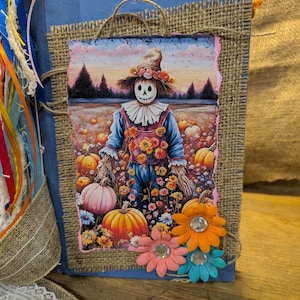 May include: A handmade journal with a burlap and blue fabric cover. The front features a colorful illustration of a scarecrow in a pumpkin patch. Embellished with decorative flowers and ribbons, this journal is perfect for fall or autumn-themed journaling.