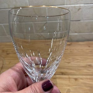 May include: A clear wine glass with a gold rim. The glass has a detailed, etched design on the lower half. The glass is held up against a light-colored background.