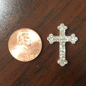 May include: A silver cross with clear rhinestones, next to a US penny for size comparison.