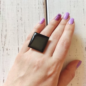 Pode incluir: Um anel quadrado preto com acabamento brilhante. O anel é usado em uma mão com esmalte de unha roxo claro.