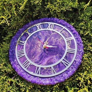 May include: A circular clock with a purple resin base and a silver clock face featuring Roman numerals. The clock face has a marbled purple and gold pattern. The outer edge is adorned with small, sparkling purple crystals. Red clock hands.