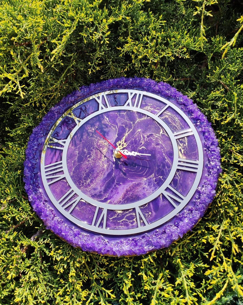 May include: A circular clock with a purple resin base and a silver clock face featuring Roman numerals. The clock face has a marbled purple and gold pattern. The outer edge is adorned with small, sparkling purple crystals. Red clock hands.