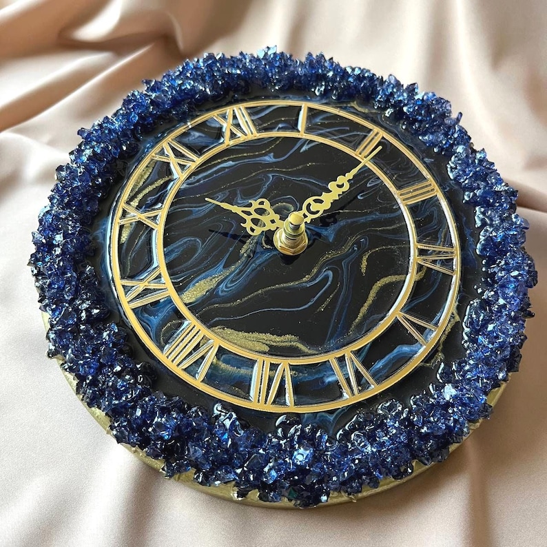 May include: A circular clock with a black and gold face, displaying Roman numerals and decorative gold hands. The clock is encircled by a border of blue crystals. The clock face has a marbled design with blue and gold detailing.