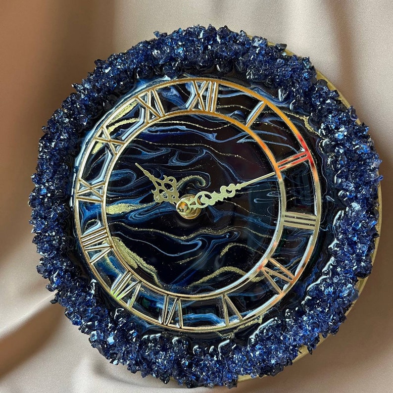 May include: A circular wall clock featuring a dark blue and black marbled resin face with gold accents. The clock has a gold-toned frame with Roman numerals and ornate gold hands. The outer edge is decorated with textured, deep blue crushed glass.