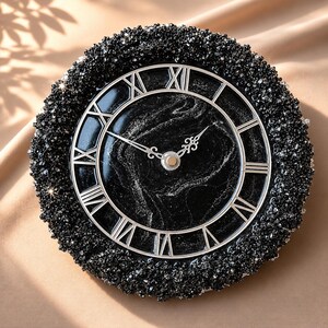 May include: A round wall clock featuring a black and silver marbled face with silver Roman numerals and ornate silver hands. The clock is framed by a thick, textured border of small, dark, reflective black crystals that catch the light.