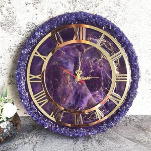May include: A circular wall clock with a purple and gold design. The clock face has Roman numerals and gold hands. The outer edge has a textured, purple border. The clock is a decorative home decor item.