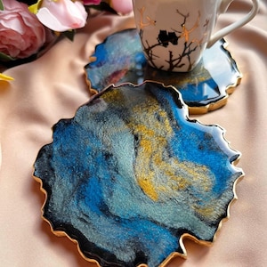 Puede incluir: Una taza de café blanca con café negro y un diseño de ramas doradas descansa sobre un posavasos de resina azul y dorada. El posavasos tiene un borde dorado y una forma orgánica. Rosas rosas en el fondo.