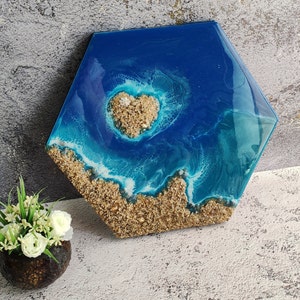 May include: Hexagon-shaped resin art piece depicting an ocean scene. The artwork features a heart-shaped island of sand surrounded by blue and turquoise resin, mimicking waves. The bottom edge is textured with sand, creating a beach effect.