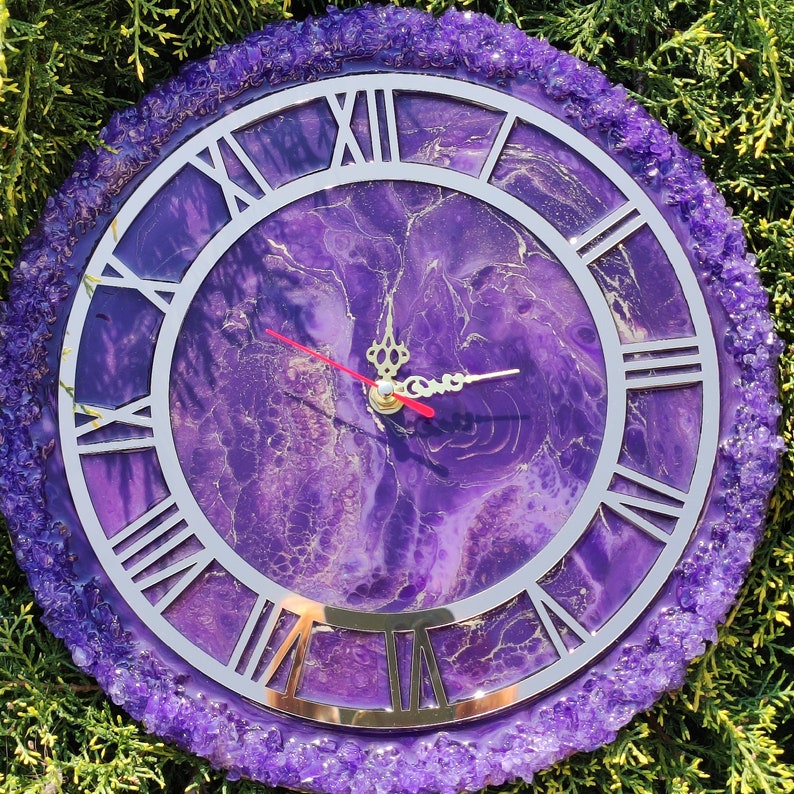 May include: A round wall clock featuring a purple and gold marbled resin face with silver Roman numerals. The clock has ornate gold hands, including a red second hand, and is bordered by a textured, crystalline purple edge.