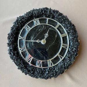 May include: A circular wall clock featuring a black and silver design. The clock face is black with silver Roman numerals and hands. The clock is encircled by a textured, dark grey border.