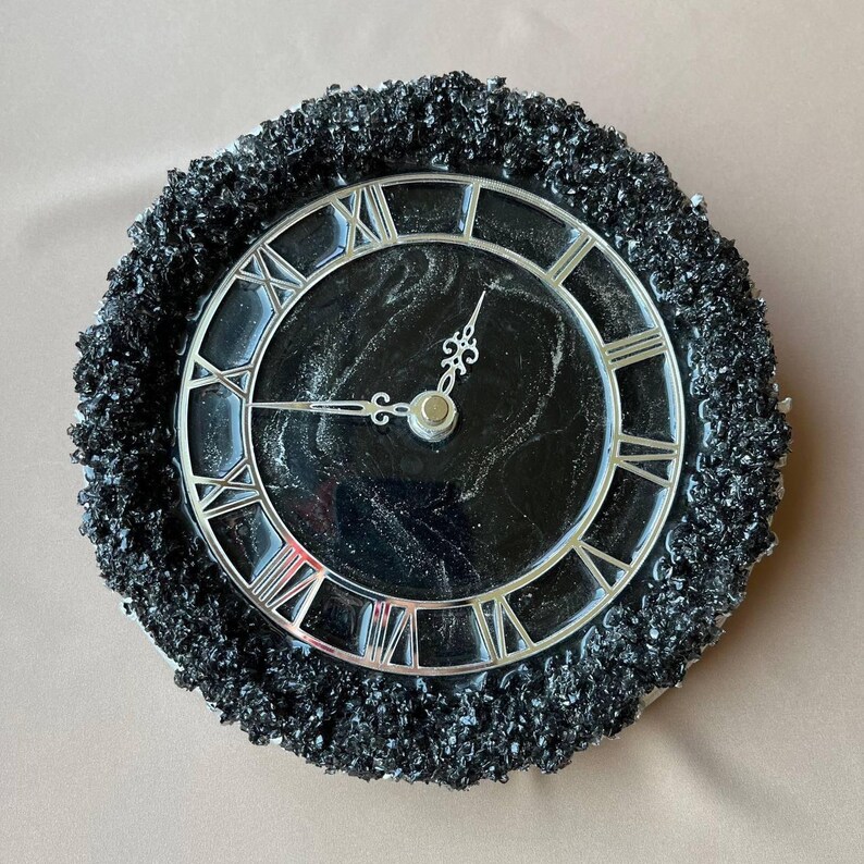 May include: A circular wall clock featuring a black and silver design. The clock face is black with silver Roman numerals and hands. The clock is encircled by a textured, dark grey border.