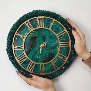 May include: A round wall clock with a gold-coloured Roman numeral face and hands. The clock face has a green and gold marbled design. The clock is surrounded by a textured, dark green border. The clock is held by two hands.