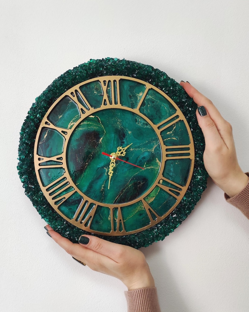 May include: A round wall clock with a gold-coloured Roman numeral face and hands. The clock face has a green and gold marbled design. The clock is surrounded by a textured, dark green border. The clock is held by two hands.