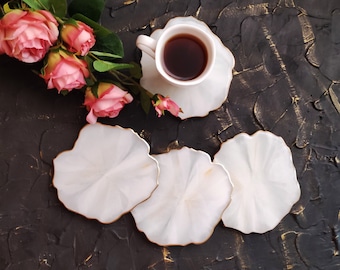 May include: Four white coasters with a gold rim and a wavy edge. The coasters are on a black surface with a cup of coffee on one of the coasters.