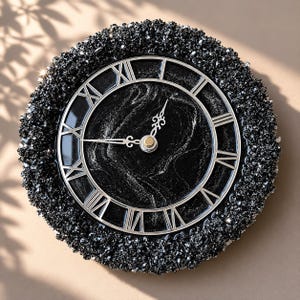 May include: A round wall clock featuring a black marbled face with silver Roman numerals and ornate silver hands. The outer border is encrusted with a thick layer of textured, sparkling black crystals, creating a high-contrast, reflective appearance.