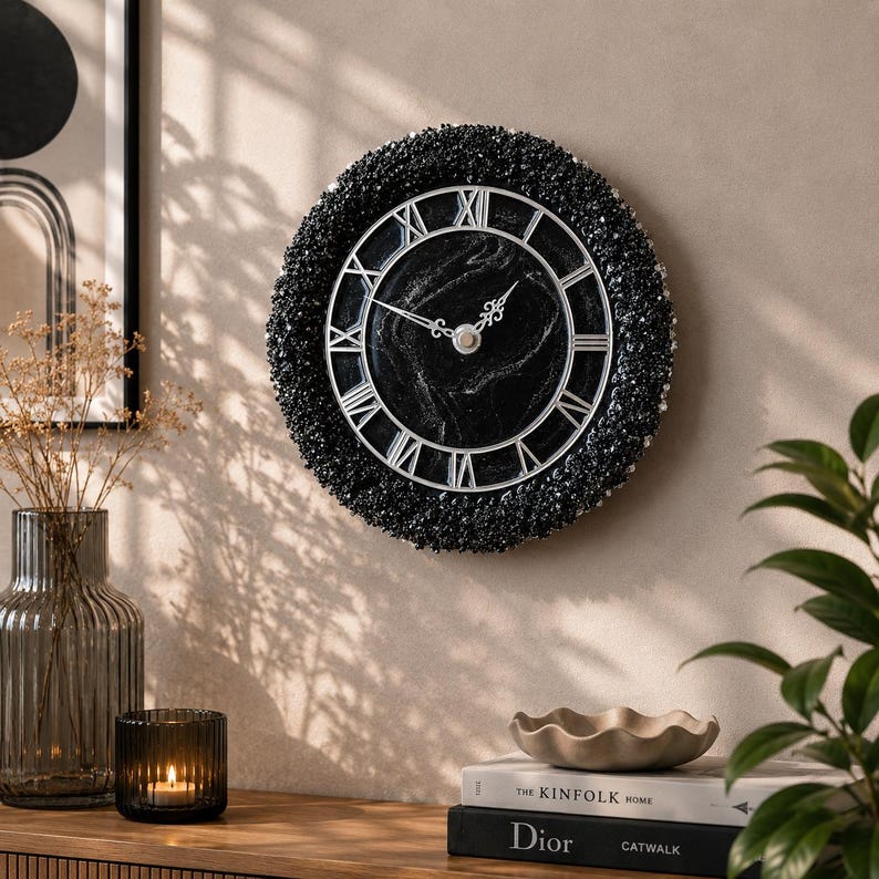 May include: A circular wall clock featuring a black marble-patterned face with silver Roman numerals and ornate hands. The clock is framed by a thick, textured border of small, dark, reflective crystals that catch the light.