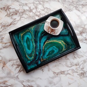 Pode incluir: Uma bandeja de servir preta com um design de resina epóxi verde e dourada. A bandeja tem um acabamento liso e brilhante e é perfeita para servir bebidas ou lanches.