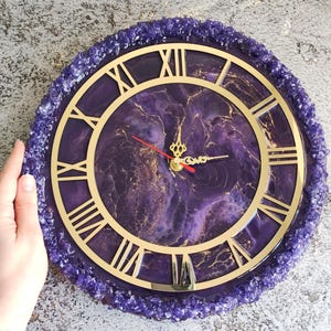May include: A round wall clock with a purple and gold design. The clock face features Roman numerals and gold hands. The clock is surrounded by a border of purple crystals. The clock's diameter is approximately 30 cm.