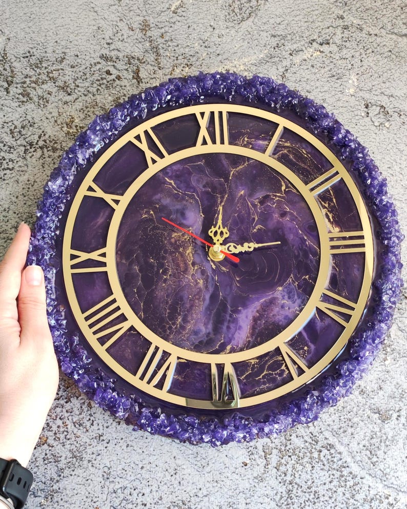 May include: A round wall clock with a purple and gold design. The clock face features Roman numerals and gold hands. The clock is surrounded by a border of purple crystals. The clock's diameter is approximately 30 cm.