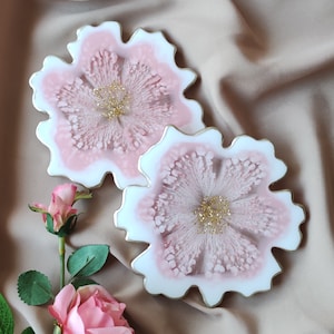 Puede incluir: Dos posavasos de resina rosa y blanca con diseño floral. Los posavasos tienen un borde dorado y están sobre un fondo de tela beige.