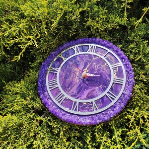 May include: A round wall clock featuring a purple and gold marbled resin face with silver Roman numerals. The outer edge is decorated with a textured border of purple crystals. The clock has silver hour and minute hands and a thin red second hand.
