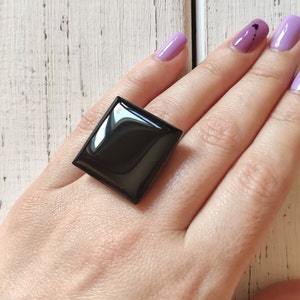 May include: A black square ring with a glossy finish. The ring is worn on a person's finger.