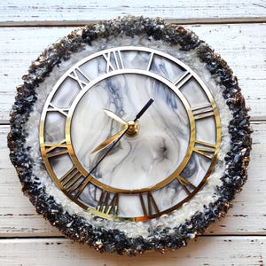 May include: Circular wall clock featuring a gold-coloured Roman numeral dial and hands. The clock face has a white and grey marble pattern. The outer edge is adorned with black and grey crystals, creating a decorative timepiece.