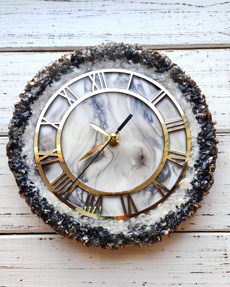 May include: Circular wall clock featuring a gold-coloured Roman numeral dial and hands. The clock face has a white and grey marble pattern. The outer edge is adorned with black and grey crystals, creating a decorative timepiece.