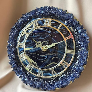 May include: Round wall clock with a dark blue and gold marbled face, silver Roman numerals, and ornate hands. The clock is surrounded by a border of dark blue crystals. The clock's face has a diameter of approximately 25 cm.