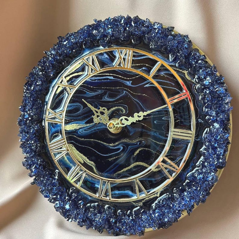 May include: Round wall clock with a dark blue and gold marbled face, silver Roman numerals, and ornate hands. The clock is surrounded by a border of dark blue crystals. The clock's face has a diameter of approximately 25 cm.