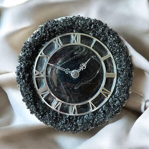 May include: A circular clock with a black face and silver Roman numerals. The clock has silver hands and a textured, dark grey outer edge. The clock is a decorative item. The clock face measures approximately 15 cm in diameter.