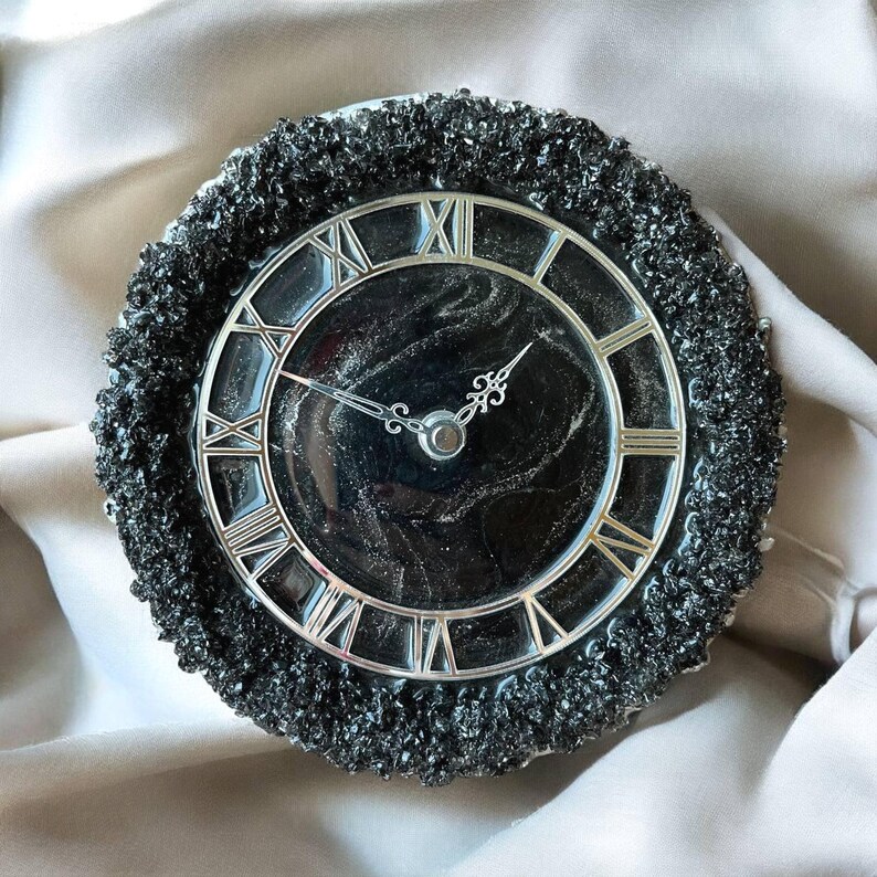 May include: A circular clock with a black face and silver Roman numerals. The clock has silver hands and a textured, dark grey outer edge. The clock is a decorative item. The clock face measures approximately 15 cm in diameter.