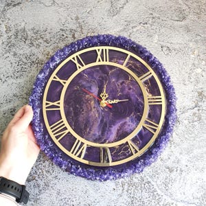 May include: A round wall clock with a purple and gold marbled face, gold Roman numerals, and a purple crystal-like border. The clock hands are red and gold. The clock's diameter is approximately 30 cm.