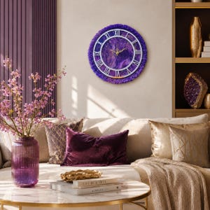 May include: A round wall clock featuring a deep purple marbled face with white Roman numerals and gold hands. The clock is framed by a textured, dark purple border. The design incorporates a circular white frame overlaying the marbled surface.