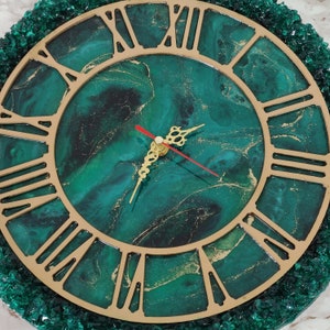 May include: A circular wall clock featuring a green and gold marbled design with gold Roman numerals. The clock hands are gold with a red second hand. The clock is framed by a border of green crystals. A decorative item for the home.