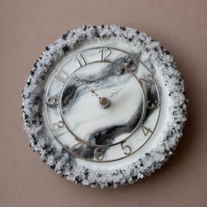 May include: A round wall clock featuring a white and black marbled face with silver-coloured numerals. The outer edge is decorated with a textured border of small, clear and black crystalline stones. It has two ornate, thin metal clock hands.