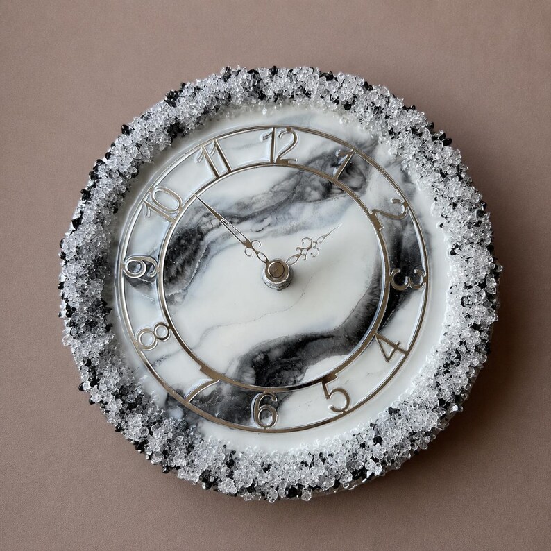 May include: A round wall clock featuring a white and black marbled face with silver-coloured numerals. The outer edge is decorated with a textured border of small, clear and black crystalline stones. It has two ornate, thin metal clock hands.