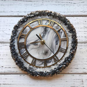 May include: Circular wall clock featuring a gold-coloured Roman numeral face and hands. The clock face has a white and grey marble design. The clock is framed by a textured, dark grey and black border. The clock is on a white wooden surface.